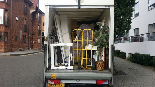 The open rear of a small commercial waste collection vehicle parked on a city street shows various discarded items intended for rubbish removal. Inside, there are several large, irregularly shaped objects, including a white plastic or foam structure with a smooth surface and rounded edges, and a tall, yellow, wheeled hand truck positioned upright beside them. To the right of the hand truck, an assortment of green plants with lush foliage is visible, some trailing down from a small basket or container. The interior also contains other miscellaneous rubbish, possibly household or garden waste, arranged in a cluttered manner. The vehicle is situated on a paved street with multi-storey residential and commercial buildings on either side, including a red brick apartment building to the left and a modern white building to the right. The scene is captured in daylight, with natural lighting highlighting the textures of the various materials and the urban environment in Pimlico, London, where Business Waste Removal Pimlico operates.