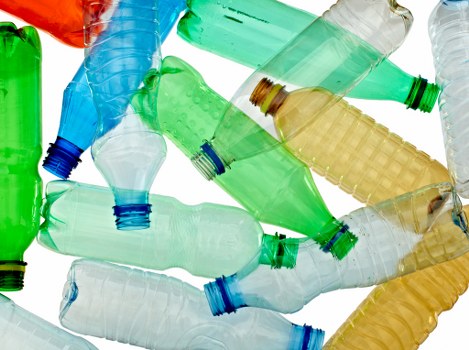 A collection of discarded plastic bottles in various colours, primarily green, blue, clear, and amber, lying haphazardly on a flat surface, with some bottles overlapping or leaning against each other. The bottles are made of lightweight PET plastic, featuring textured grips and smooth, glossy surfaces that reflect light. The open necks of the bottles are visible, and some have slight creases or deformation from use. The scene suggests a waste collection area or a site for rubbish removal, with the bottles scattered in a seemingly random arrangement. The background is plain and neutral, emphasizing the bottles' vivid colours and varied textures. This image illustrates typical clutter associated with plastic waste and aligns with professional rubbish removal services like those offered by Business Waste Removal Pimlico, situated in central London, serving local businesses that generate plastic packaging waste. The overall lighting highlights the transparency and glossy finish of the plastic bottles, reinforcing the focus on waste collection and disposal processes common in urban waste management in the Pimlico area.