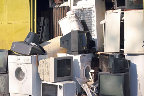 A collection of discarded electronic waste and household appliances piled together outdoors, including various models of washing machines, microwaves, televisions, and computer monitors. The appliances show different textures and finishes, such as white, black, and grey plastic casings with metallic components, and some screens displaying dark or reflective surfaces. The objects are arranged in a haphazard manner, with some stacked on top of each other and others leaning against the surrounding items. In the background, a metal and wooden structure is partially visible, suggesting an outdoor storage or waste collection area. Natural daylight illuminates the scene, emphasizing the varied surfaces and textures of the waste items. This image exemplifies typical bulky rubbish that a professional waste removal service like Business Waste Removal Pimlico would handle, especially in urban environments near Pimlico, London, where such waste collection is common.