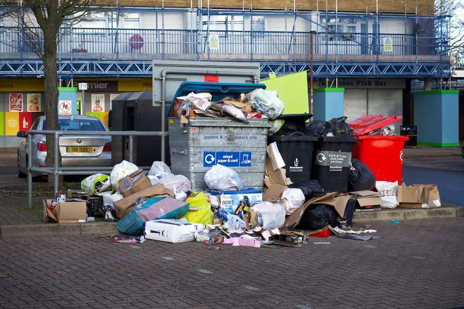 The image shows a collection of overflowing rubbish and waste bags placed around a large grey communal trash bin on a paved outdoor area. The bin is filled with mixed waste paper, cardboard, and plast