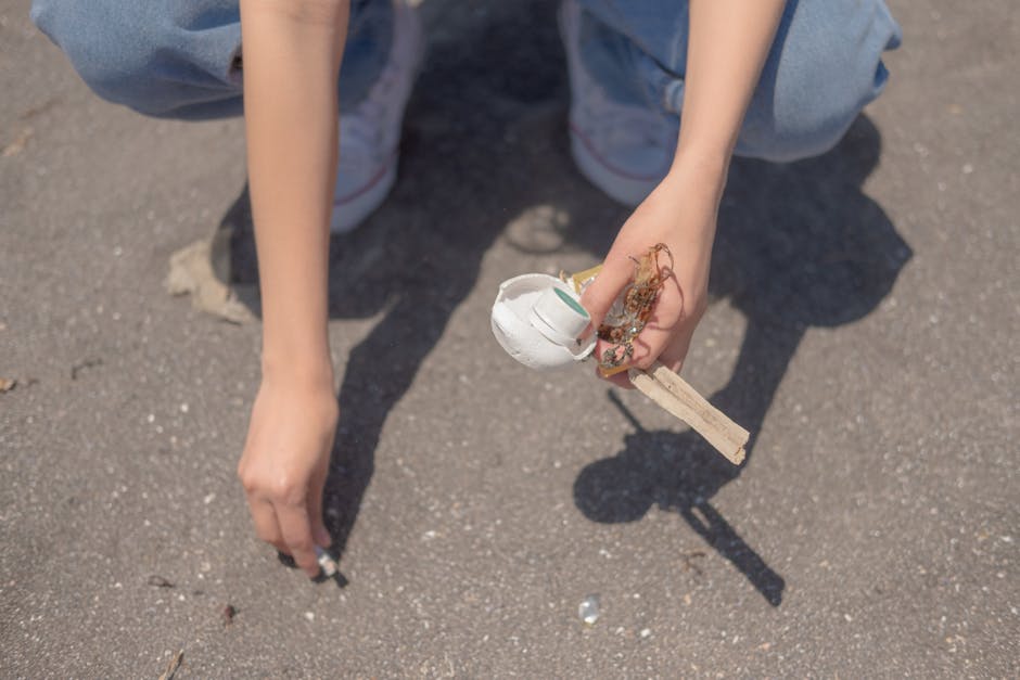 An overflowing collection of mixed waste and recyclable rubbish is piled on a paved sidewalk beside a metal railing, including cardboard boxes, plastic bags, paper, and packaging materials. The waste 